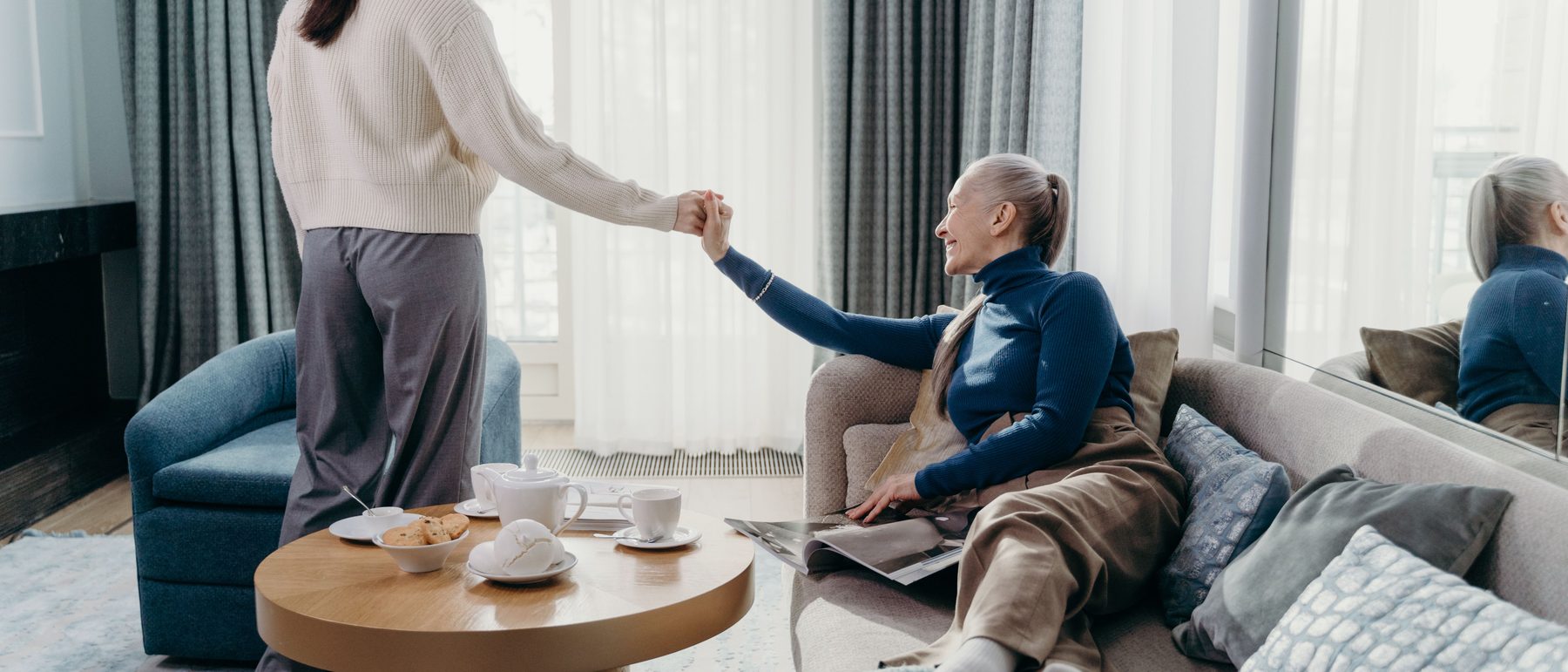 A woman reaching out to hold the hand of an older woman relaxing on a sofa in a bright living room